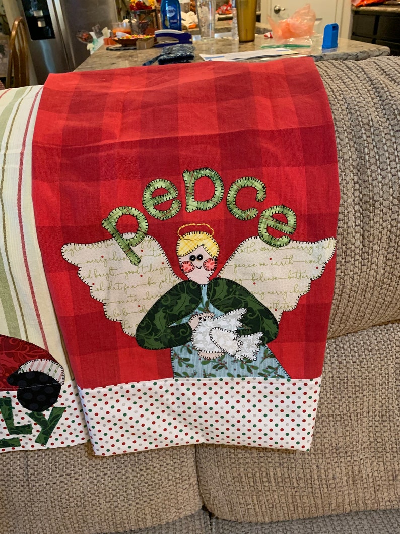 May include: Red and white checkered kitchen towel with a green and white angel holding a white dove. The angel has blonde hair and blue eyes. The towel has the word "peace" embroidered in green on it.