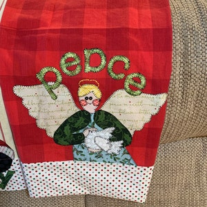 May include: Red and white checkered kitchen towel with a green and white angel holding a white dove. The angel has a halo and the word "peace" is spelled out in green letters above the angel.