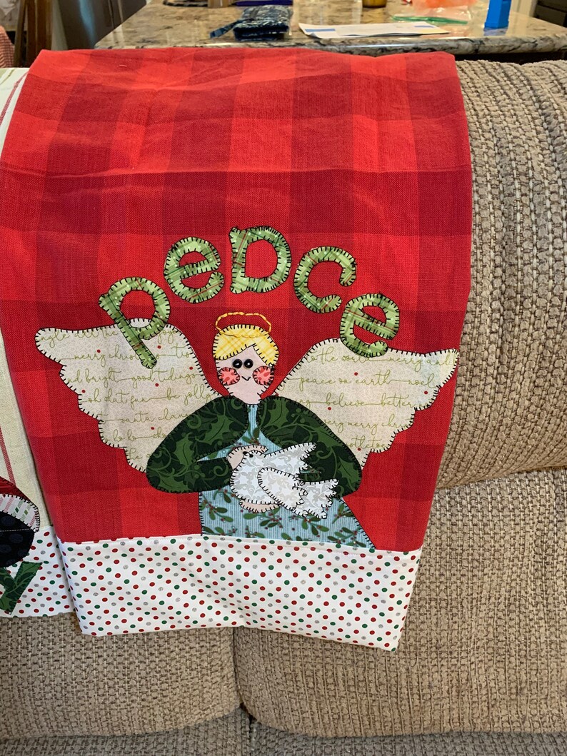 May include: Red and white checkered kitchen towel with a green and white angel holding a white dove. The angel has a halo and the word "peace" is spelled out in green letters above the angel.