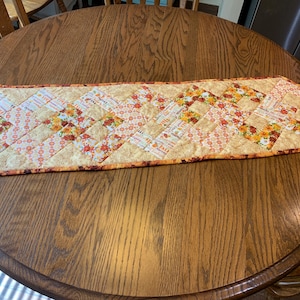May include: A quilted table runner with a fall theme. The runner is made of various fabrics with floral and geometric patterns in shades of orange, yellow, and brown. The runner has a scalloped edge.