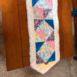 May include: A colorful quilted table runner with a sunflower design at each end. The runner features blue, pink, and floral patterned fabric squares. The edges are trimmed with white fabric.