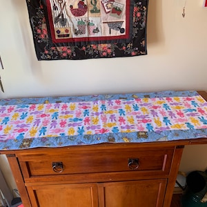 May include: A blue and white table runner with a pattern of colorful bunnies. The runner is on a wooden table with a drawer and a cabinet.