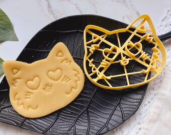 Puede incluir: Un cortador de galletas de plástico amarillo en forma de cara de gato con corazones para los ojos. Se muestra una galleta cortada con el cortador junto a él.