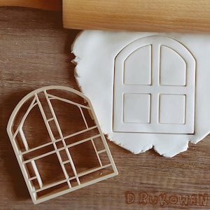 May include: A white plastic cookie cutter shaped like a window with a rounded top and six panes. The cookie cutter is on a wooden surface with a rolling pin and a piece of white fondant.