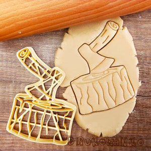 May include: Two gold-coloured cookie cutters in the shape of an axe stuck in a log. The cutters are on top of a sheet of unbaked cookie dough.