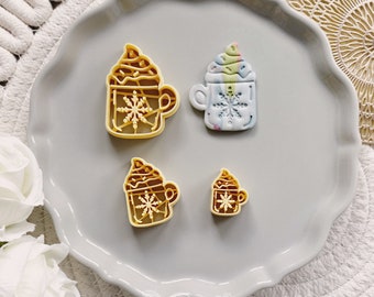 May include: Four cookie cutters in the shape of a winter-themed hot chocolate drink. The cutters are made of yellow plastic and feature a snowflake design. The cutters are arranged on a gray plate.