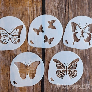 Peut inclure: Cinq pochoirs en plastique blanc avec des motifs de papillons. Les pochoirs sont circulaires et présentent différentes formes et tailles de papillons.