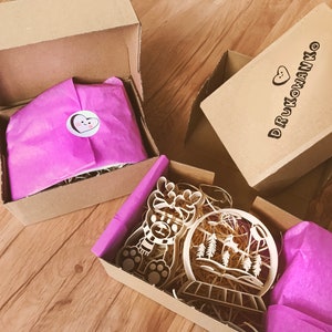Op de afbeelding: Drie kartonnen dozen met roze tissuepapier en houten uitsnijdingen. De dozen zijn gelabeld met "DRUKOWANKO" met een hartvormig ontwerp. De houten uitsnijdingen zijn een rendier, een sneeuwbol en een set poot afdrukken.