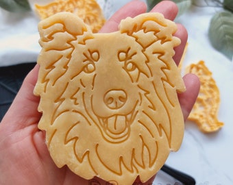 May include: A light brown cookie cutter in the shape of a dog's head, held in a hand. The dog has detailed facial features, including eyes, a nose, and a smiling mouth. The cookie cutter is surrounded by other similar cutters.
