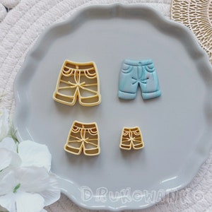 Puede incluir: Cuatro cortadores de galletas con forma de pantalones cortos. Dos son amarillos y dos son azules. Los cortadores amarillos son de diferentes tamaños. Los cortadores azules son del mismo tamaño y tienen un diseño de mármol.