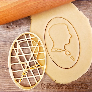 May include: A close-up of a cookie cutter in the shape of a person's head. The cutter is made of plastic and has a grid pattern. The cutter is sitting on a piece of dough.