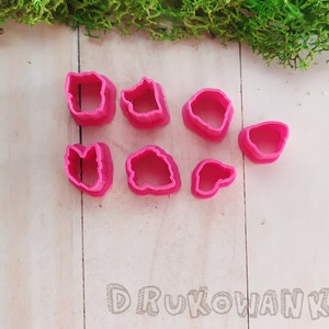 Op de afbeelding: Een set van acht roze plastic koekjesvormen in verschillende vormen, waaronder bladeren, bloemen en harten. De vormen zijn gerangschikt op een wit houten oppervlak.