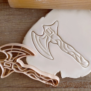 May include: A white plastic cookie cutter in the shape of an axe with a detailed design. The cutter is on a white surface.