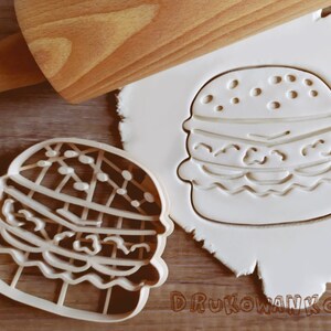 May include: Two white plastic cookie cutters shaped like hamburgers. The cutters are on a wooden surface. The cutter on the left is detailed with sesame seeds, lettuce, and tomato. The cutter on the right is a more basic design.