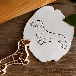 May include: A white dachshund cookie cutter on a white background. The cookie cutter is made of metal and has a detailed design. There is a second cookie cutter in the shape of a dachshund on a wooden surface.
