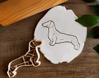 May include: A white dachshund cookie cutter on a white background. The cookie cutter is made of metal and has a detailed design. There is a second cookie cutter in the shape of a dachshund on a wooden surface.