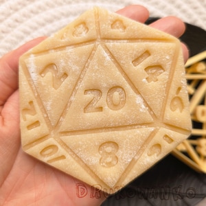 Puede incluir: Un primer plano de una galleta hecha en casa con forma de dado de veinte caras. La galleta está espolvoreada con azúcar en polvo y tiene los números 2, 12, 14, 6, 10, 8 y 20 impresos en ella.