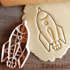 May include: A close-up of a cookie cutter in the shape of a rocket ship. The cookie cutter is made of metal and has a detailed design. The cookie cutter is on a wooden surface next to a piece of cookie dough.
