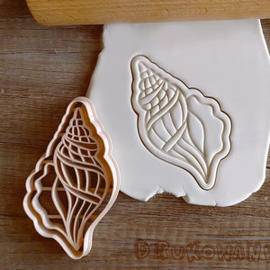 Puede incluir: Un cortador de galletas de plástico con forma de concha. El cortador está detallado con líneas y curvas intrincadas. El cortador se muestra sobre una superficie blanca.