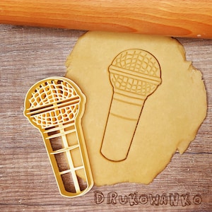 Pode incluir: Um cortador de biscoitos dourado em forma de microfone. O cortador está sobre uma superfície marrom com um rolo de massa no fundo.