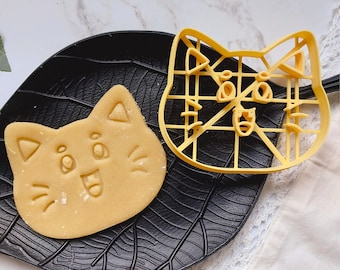 Puede incluir: Un cortador de galletas de plástico amarillo en forma de cara de gato con un patrón de cuadrícula. Una galleta en forma de cara de gato está en un plato negro con forma de hoja.