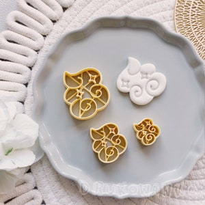 Puede incluir: Cuatro cortadores de galletas en forma de nubes con estrellas. Los cortadores están hechos de plástico de color dorado y están dispuestos en un plato gris.