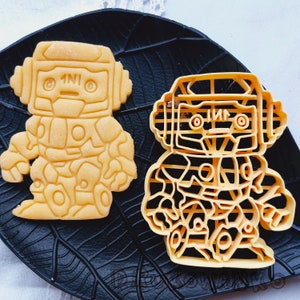 Puede incluir: Dos cortadores de galletas amarillos con forma de robots. El robot de la izquierda tiene una cabeza redonda con un solo ojo a cada lado y el texto "1N10" en la frente. El robot de la derecha tiene un diseño más detallado con una cabeza rectangular y un solo ojo a cada lado. El texto "1N10" también está en la frente de este robot.
