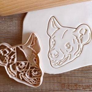 May include: A white plastic cookie cutter in the shape of a hyena head. The cutter is on a wooden surface.