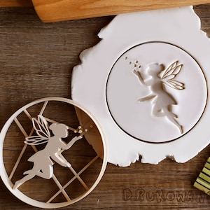 Puede incluir: Un cortador de galletas de plástico blanco con un diseño de hada soplando burbujas. El cortador se muestra junto a un rodillo de madera y una estera de bambú.