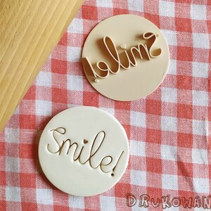 May include: Two white round cookie cutters with the words "Smile!" and "Hello" on them. The cookie cutters are on a red and white checkered tablecloth.
