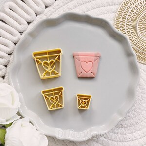 Può includere: Quattro formine per biscotti a forma di vaso, ciascuna con un design a cuore. Due sono gialle e due rosa. Le formine sono disposte su un piatto grigio con bordo smerlato, con uno sfondo strutturato bianco e grigio.