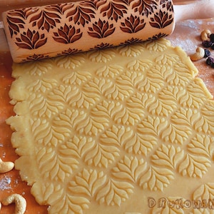 May include: A wooden rolling pin with a carved floral design is used to roll out dough, creating a patterned surface on the dough. The dough is light brown and the rolling pin is brown with a natural wood finish.