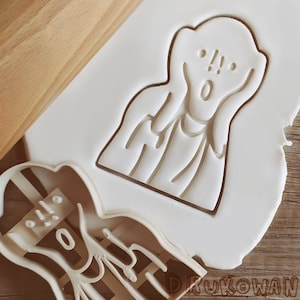 Puede incluir: Un cortador de galletas blanco con la forma de la obra "El Grito" de Edvard Munch, junto a una galleta cortada y un rodillo. El cortador de galletas tiene los rasgos de la cara claramente definidos. La galleta cortada está sobre una superficie blanca.