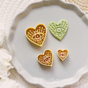 Puede incluir: Cuatro cortadores de galletas en forma de corazón, dos son amarillos y dos son verdes. Los cortadores presentan un ojo con una luna creciente y un sol encima, rodeado de un patrón geométrico.
