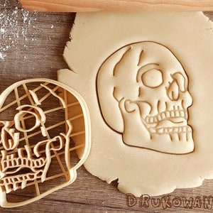 May include: A close-up of a cookie cutter in the shape of a skull. The cookie cutter is made of metal and has a detailed design. The cookie cutter is sitting on a piece of dough.