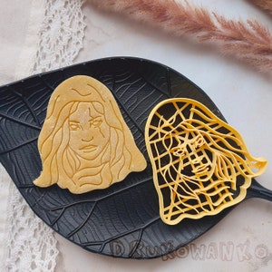 May include: Two yellow cookie cutters shaped like women's faces with long hair. The cutters are on a black leaf-shaped plate.