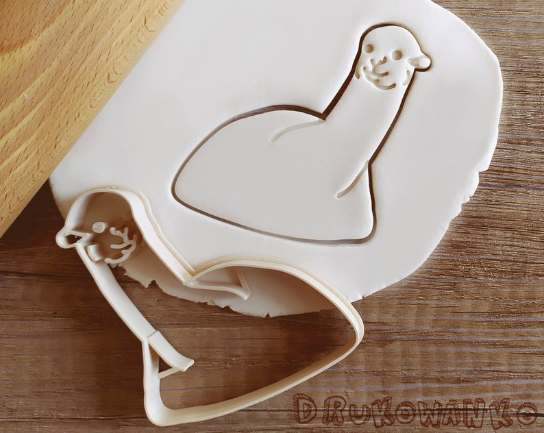 Pode incluir: Um cortador de biscoitos de pl&aacute;stico branco em forma de lhama de desenho animado. O cortador est&aacute; sobre uma superf&iacute;cie branca com um rolo de massa no fundo.