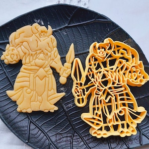 May include: Two cookie cutters, one shaped like a knight with a sword and the other shaped like a warrior with a shield and sword. Both are made of yellow metal and are on a black background.
