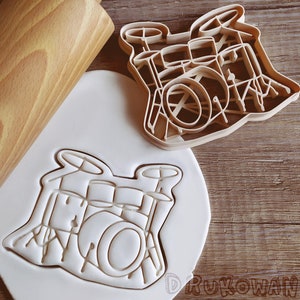 May include: A white plastic cookie cutter in the shape of a drum set. The cookie cutter is on a white surface with a wooden rolling pin in the background.
