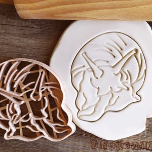 May include: A white plastic cookie cutter in the shape of a curled up snail. The cutter is shown on a wooden surface.