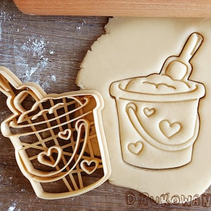May include: Two cookie cutters, one shaped like a bucket with a shovel and the other shaped like a bucket with a straw. Both cutters have hearts on them.