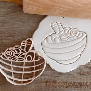 May include: Two white plastic cookie cutters shaped like bowls of ramen noodles. The cutter on the left is detailed with a chopsticks and noodles. The cutter on the right is a more simple design with a bowl of noodles.