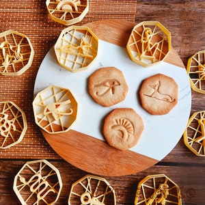May include: A set of 10 cookie cutters in various shapes, including an ankh, a cat, and a sun. The cutters are made of a light yellow plastic and are arranged on a wooden surface.