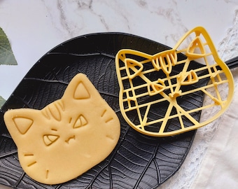 Puede incluir: Dos cortadores de galletas de plástico amarillo, uno con forma de cara de gato con una expresión de enfado y el otro con forma de cabeza de gato con un diseño geométrico. Los cortadores están sobre una superficie negra.