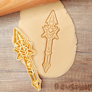 Puede incluir: Dos cortadores de galletas de color dorado en forma de lanzas. Los cortadores están sobre una superficie de madera marrón con un rodillo y una masa.