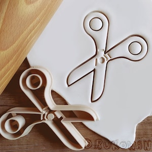 Può includere: Un tagliapasta in plastica bianca a forma di forbici. Il tagliapasta è su una superficie bianca. Un mattarello è sullo sfondo.