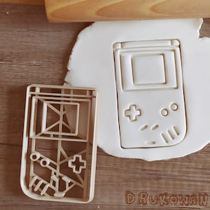 May include: A white plastic cookie cutter shaped like a handheld video game console. The cookie cutter has a screen, buttons, and a directional pad.