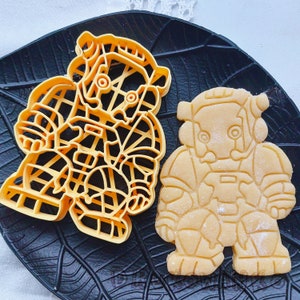 Puede incluir: Dos cortadores de galletas, uno con forma de robot y el otro con forma de persona. El cortador de galletas de robot está hecho de una masa marrón claro y el cortador de galletas de persona está hecho de una masa amarilla.