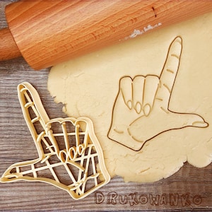 May include: Two cookie cutters in the shape of hands making the 'L' sign. The cutters are on a sheet of cookie dough. The cookie cutters are made of metal and have a shiny finish.