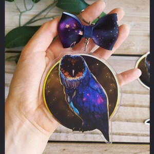 May include: A clear acrylic charm with a galaxy owl design. The owl is dark blue and purple with a yellow background. The charm is attached to a black and white bow.
